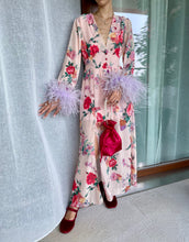 Carica l'immagine nel visualizzatore di Gallery, Abito maxi manica lunga elasticizzato in vita e bottoncini stile corsetto. Fantasia rosa pesca floreale, con boa in piuma di struzzo lilla ai polsi. Spacco laterale. Attaccatura del boa tramite asoline e bottoncini cuciti a mano, per facilitare il lavaggio e per poterlo utilizzare anche senza piuma. Composizione tessuto: 50% polyester; 45% viscose; 5% elastam.